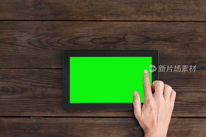 手指按在绿色屏幕的平板电脑上。俯视图在一张深棕色的木桌上。