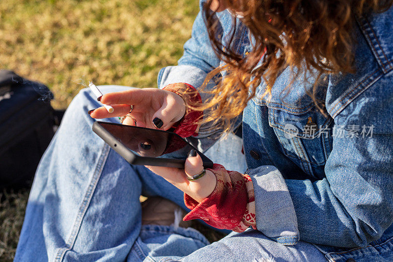 一个女人坐在公园的草地上，一边看手机一边抽烟。