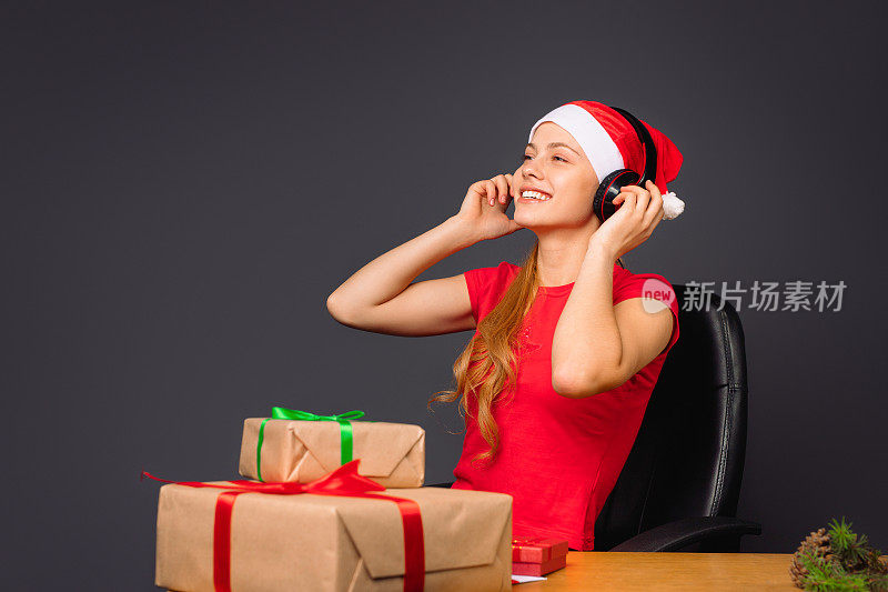 快乐的小精灵戴着大大的耳机坐在椅子上听音乐，微笑着，靠近圣诞礼物。灰色的背景。节日的概念。