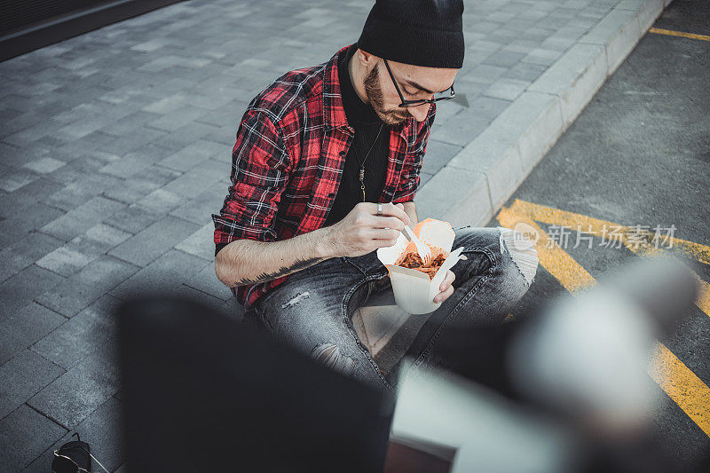 一个赶时髦的年轻人骑着电动滑板车坐在滑板车旁边休息，吃着美味的意大利面。他坐在人行道上，在商场的人行道上。