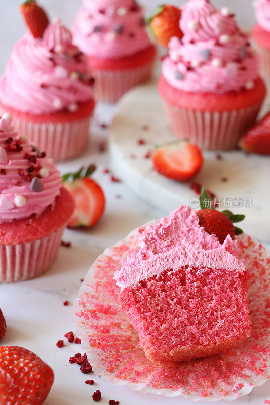 个人自制的粉色丝绒草莓纸杯蛋糕，切成两半，周围是纸制蛋糕盒里的松饼，草莓粉奶油糖霜螺旋形，糖精心形和球形点缀，注意前景