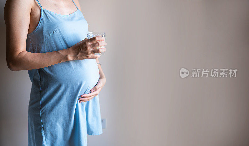 一个孕妇抱着她的肚子和一杯水在卧室靠墙
