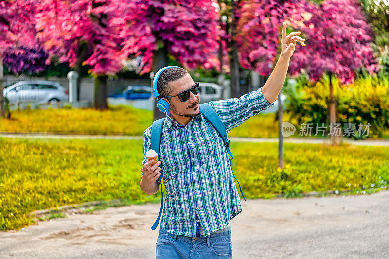 一位穿着格子衬衫的帅哥正走在城市公园里，一边听着音乐，一边吃着冰淇淋提神，一边用手臂问候别人。