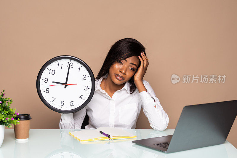 困倦的非裔美国女商人坐在桌子旁，时钟已经晚了，电脑在办公室孤立的灰色背景