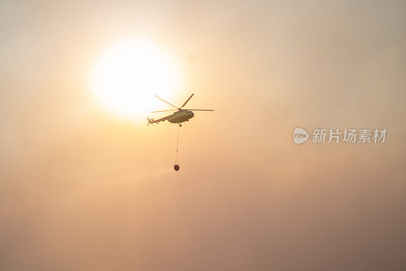 空中消防在黄色多云的天空背景中带着水篮的飞行直升机。