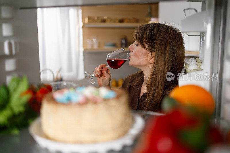 一个年轻的女人站在厨房打开的冰箱旁喝着红酒