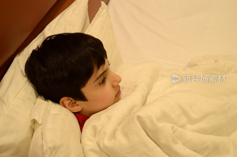一个可爱的可爱的九岁男孩躺在床上，他的头靠枕头支撑着，白色的床单和羽绒被或被子盖在他的侧面