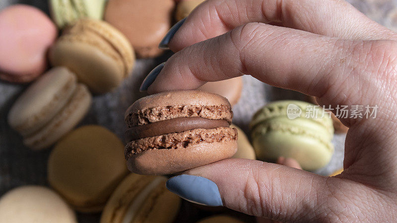 特写图像，一个难以辨认的人拿着巧克力马卡龙之间的手指和拇指在一堆多色蛋白馅奶油，香草，柠檬，咸焦糖，巧克力，草莓和开心果味道，提高的看法