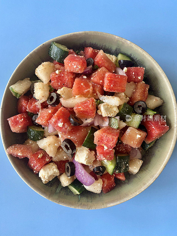 西瓜沙拉的碗，切片黄瓜，红洋葱，羊乳酪块，薄荷叶和黑橄榄，蓝色的背景，上升的观点