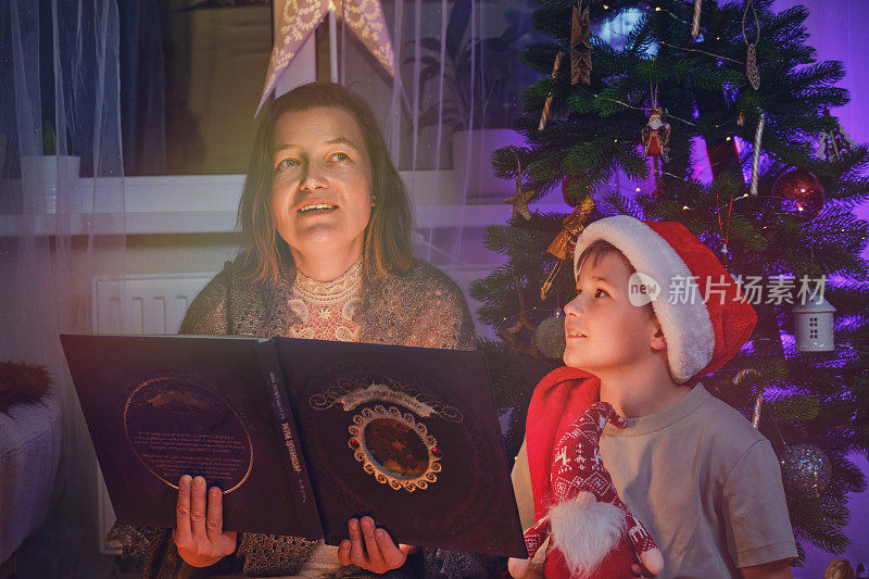 一名成年女性和一名男孩正在家庭客厅的圣诞树旁读书。妈妈和儿子在新年前夕读童话故事