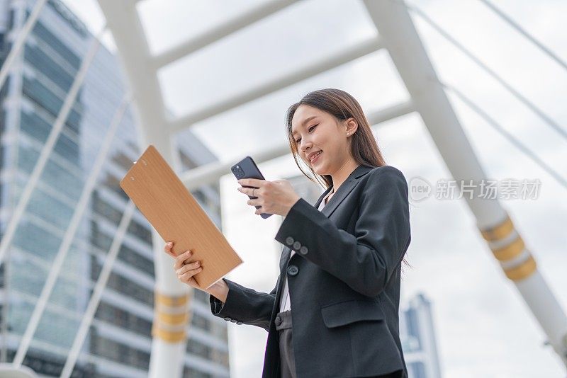 迷人的年轻亚洲职业女商人女士与一个美好的微笑夹夹板看着电话检查市场和销售库存报告在曼谷市的中央商务区