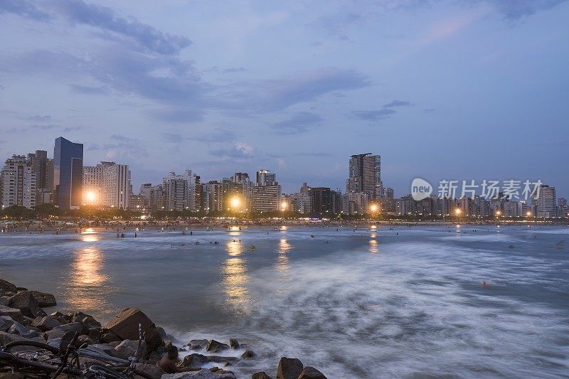 巴西桑托斯市。何塞·梅尼诺海滩。下午晚些时候，海滨建筑和拥挤的海滩和冲浪者在水中冲浪。长曝光摄影。