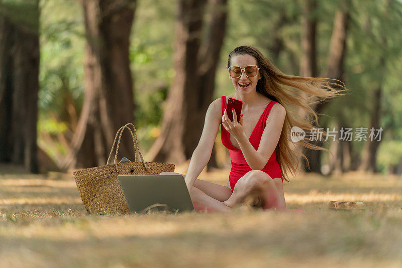 美丽的白人女性性感的女人在红色泳装比基尼太阳镜工作在她的电脑笔记本电脑和视频通话和自拍在她的手机，而坐在海边的树林放松