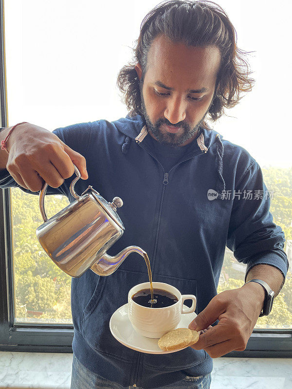 特写画面:站在酒店房间窗口前的印度男子将刚煮好的黑咖啡从不锈钢咖啡壶中倒入白色的杯子和碟子中，并配上两块饼干，聚焦前景
