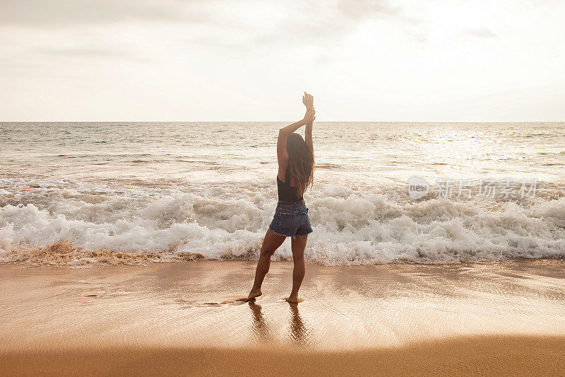 后视图年轻女子在牛仔短裤摆姿势在沙滩在海浪背景，暑假。后视图女士站在热带海岸，生活方式。旅游度假概念。复制文本空格