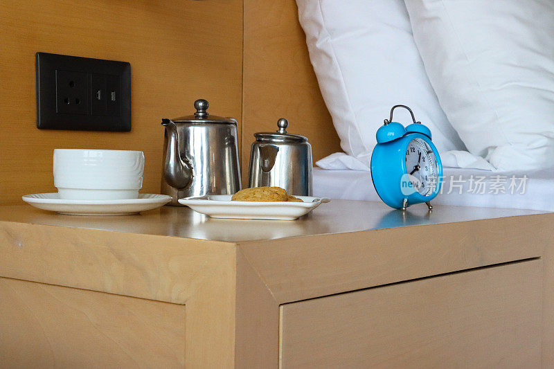 床头柜全画框形象蓝色金属外壳，复古闹钟，双钟，模拟显示时钟，白色咖啡杯碟，饼干盘，不锈钢咖啡和牛奶壶，带白色床单和枕头的双人床，电源插座