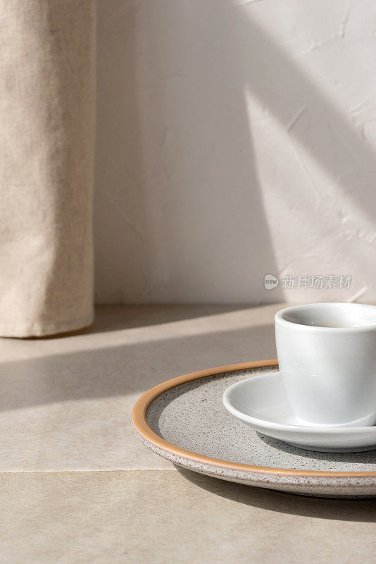 米色石桌上咖啡杯、盘子的极简构图，中性墙面上亚麻窗帘的唯美阳光阴影，清晨咖啡概念，复制空间