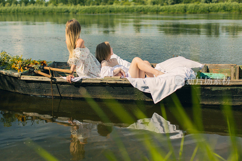 两个穿白衣服的金发女人在湖边度假。女性的友谊。旧渔船上的白色亚麻布和桨。床单。夏天放松