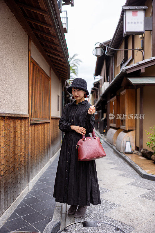 身穿黑色连衣裙、头戴帽子的年轻女子站在日本传统的狭窄街道上的全身画像