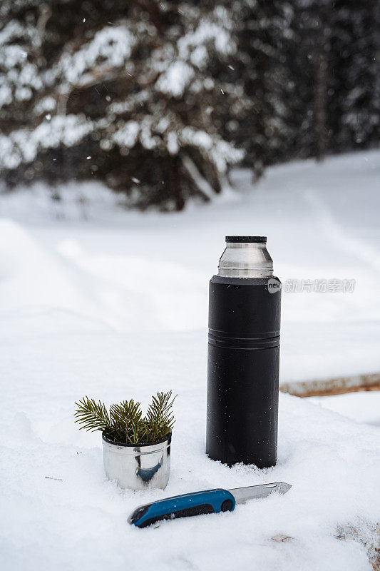 黑色哑光保温瓶，杯子上有雪中的云杉树枝和小刀。远足和野餐的工作人员。在冬天的霜冻里喝着热茶。极简主体拍摄