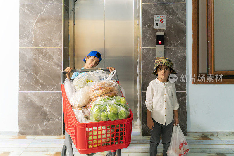 购物归来的男孩推着满满的购物车在电梯前摆姿势。