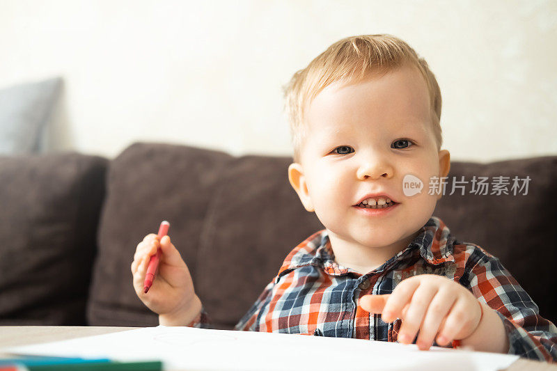 一个金发小男孩坐在桌子旁用彩色马克笔画画，看着镜头，面带微笑。