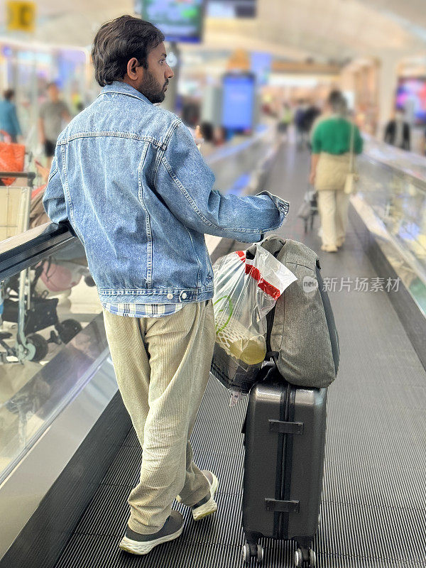 机场候机楼，印度旅客拿着飞机行李站在扶梯上旅行，手提行李堆在轮式行李箱上，焦点在前景，后视图