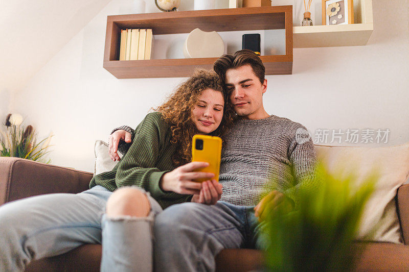 美丽的年轻夫妇在客厅放松，同时看媒体在他们的智能手机