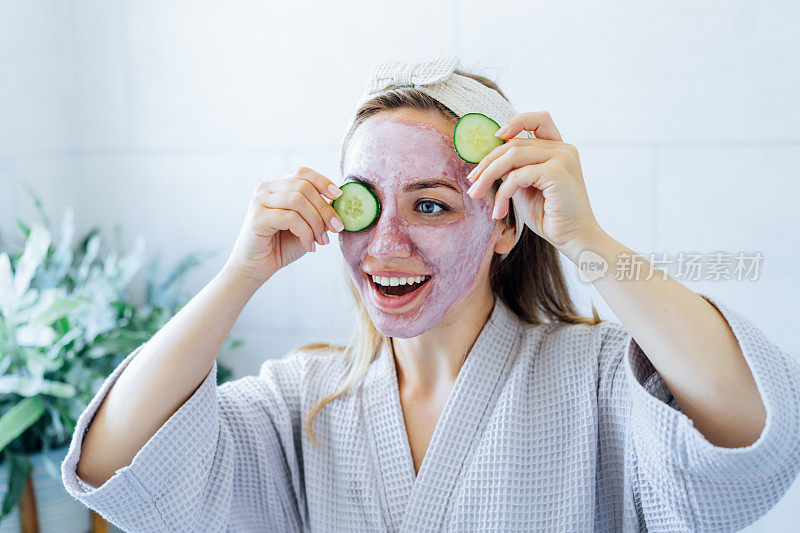 一名年轻的女子面带微笑，敷着粉色粘土面膜，手里拿着黄瓜片，在浴室里做着清爽的眼膜。自然美容程序的皮肤护理在家里。美容保健。有选择性的重点。