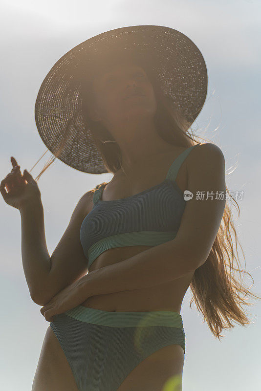 美丽的白人妇女在蓝色比基尼黑色沙滩帽和太阳镜在海滩上强烈的太阳背光穿过她的头