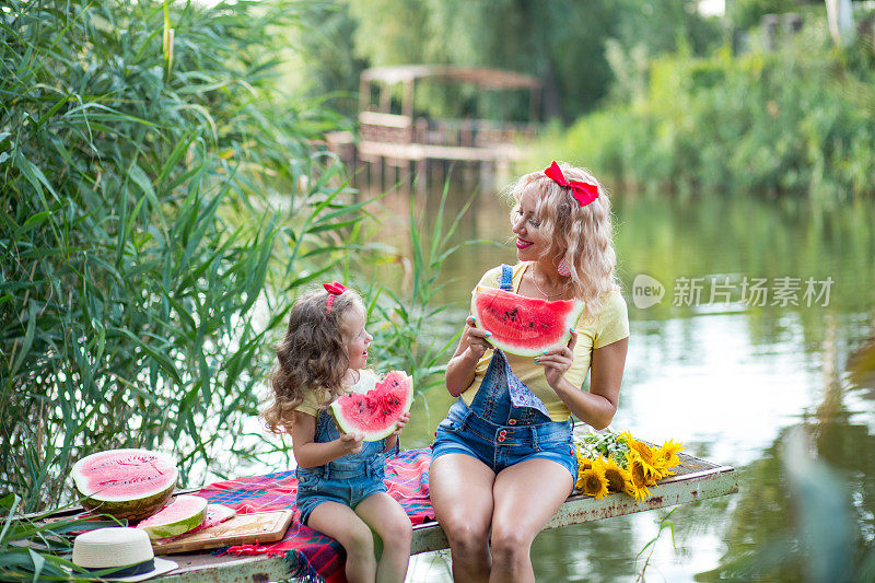 金发碧眼的母亲带着三岁的女儿，穿着牛仔短裤，系着红色的蝴蝶结，坐在码头的格子毯子上吃西瓜，河边有一束盛开的向日葵