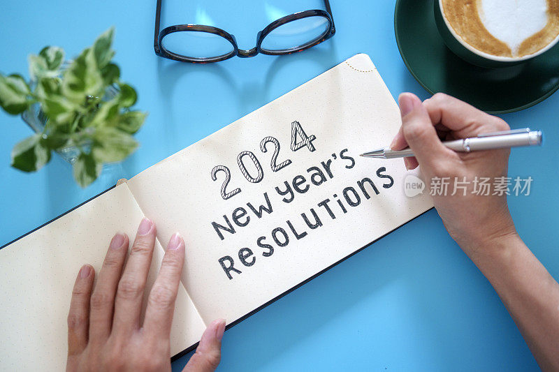 一名女性的手在她的工作桌上的纸质笔记本上写下了2024年的新年决心。