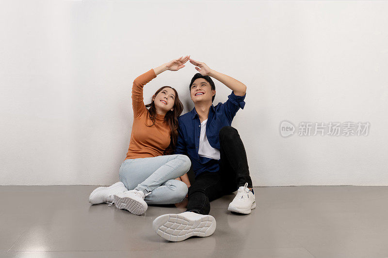 快乐的年轻亚洲夫妇穿着休闲服装坐在地板上，用手搭屋顶，搬迁的概念