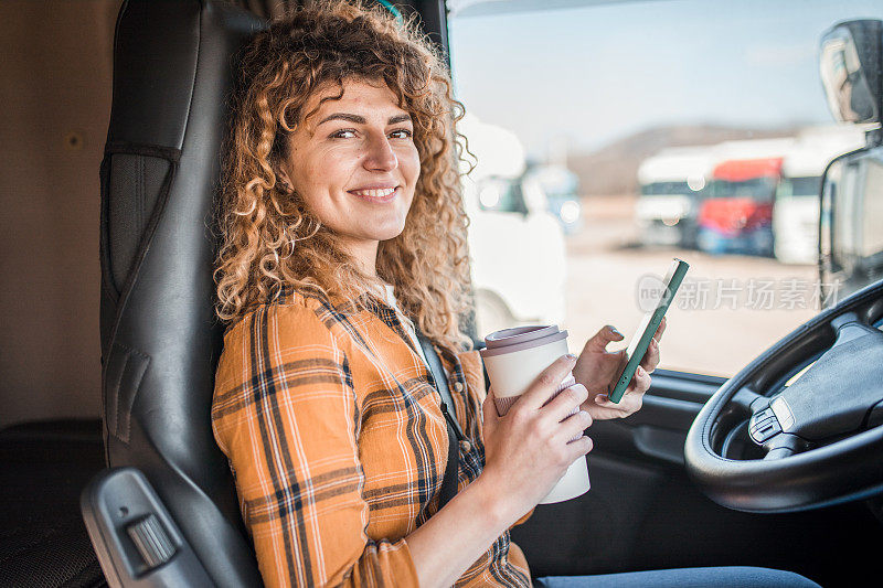 一名女卡车司机坐在卡车上，一边喝着咖啡，一边用手机，直视着镜头