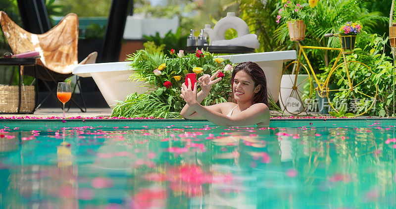 印度年轻的z世代女性站在游泳池里用手机进行视频通话。快乐的微笑美丽的放松女性自拍照片视频博主流媒体在线直播记录拍摄视频博主享受暑假豪华度假之旅