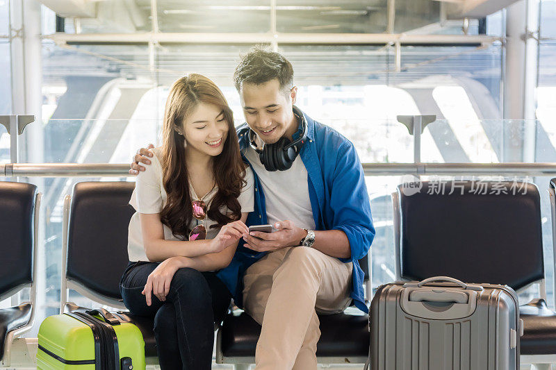 幸福亚洲夫妇带着旅行箱使用智能手机在现代机场旅行，带着科技的交通理念。