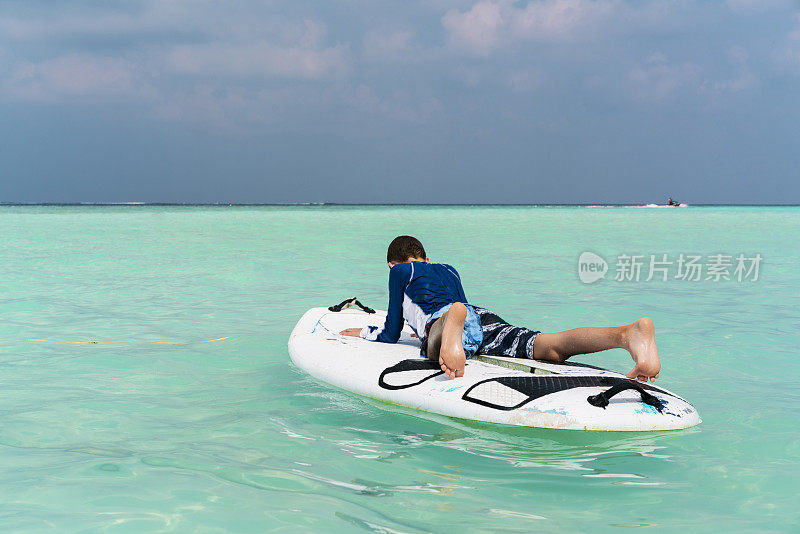 马尔代夫古利岛的比基尼沙滩上，一个小男孩躺在冲浪板上，背靠蓝天