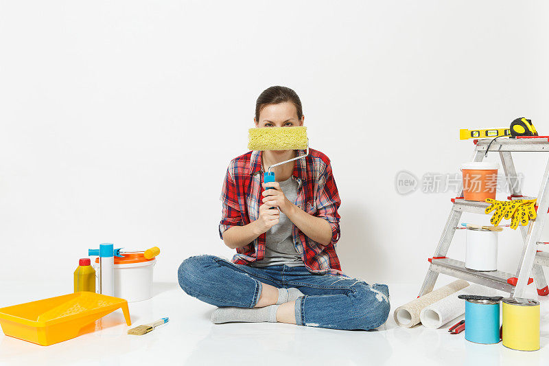 穿着便服的女人坐在地板上，用油漆滚筒，仪器为装修公寓孤立的白色背景。墙纸，胶水配件，油漆工具。修理家里的概念。