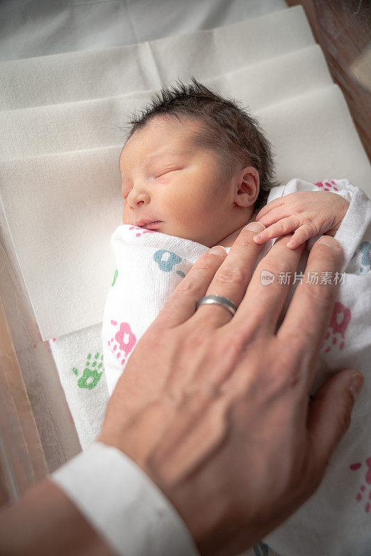 一个刚出生的女婴躺在毛毯襁褓里，她的小手和手指放在父亲布满皱纹的手上，静静地睡着了。