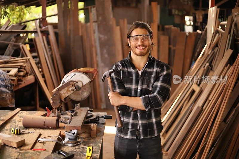 微笑的年轻白人木匠的肖像是在他自己的工匠车库风格的车间为爱好工作