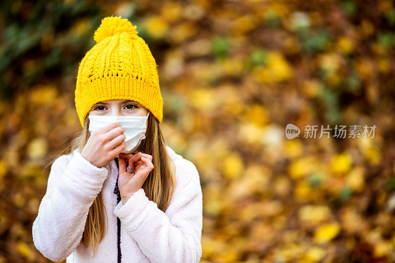 在冠状病毒感染的秋季森林里，一名小女孩戴着黄色针织帽和防护口罩