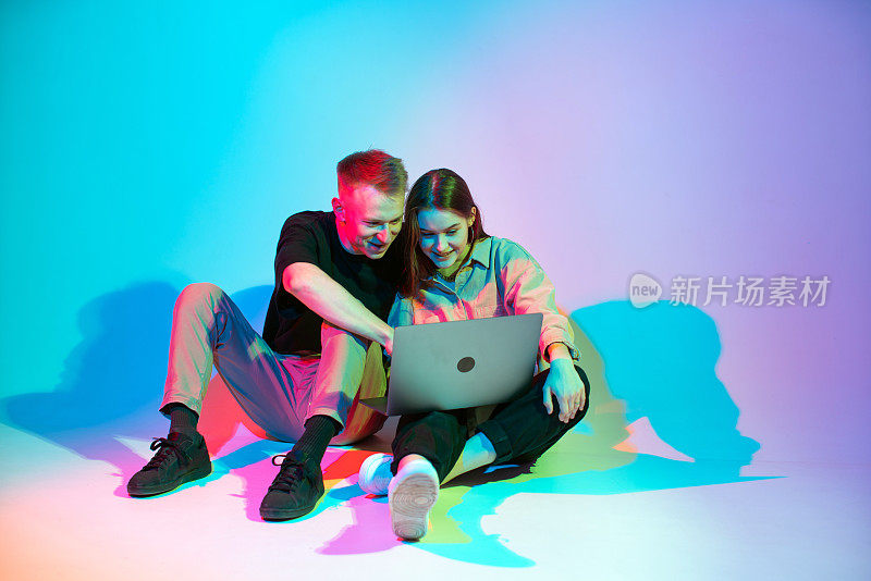 一对年轻的情侣，一个在笔记本电脑上工作，一个在网上购物。彩色霓虹灯工作室灯