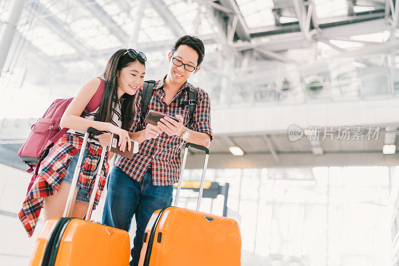 亚洲夫妇旅行者使用智能手机在机场检查航班或在线值机，带着护照和行李。航空旅行或移动电话技术概念