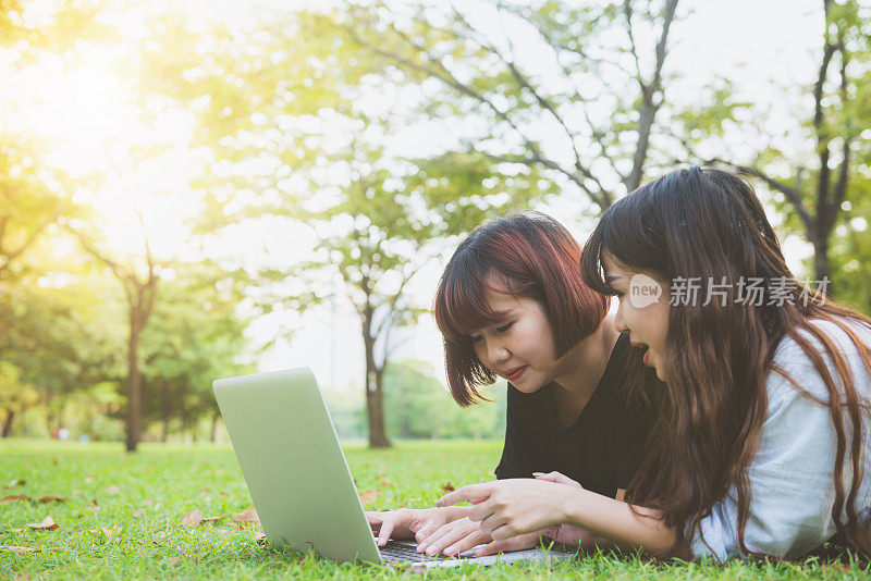 年轻的亚洲女性躺在草地上，用笔记本电脑打字。女孩的手放在键盘上。远程学习的概念。在公园里用笔记本电脑工作的快乐时髦的年轻亚洲女性。学习在户外。