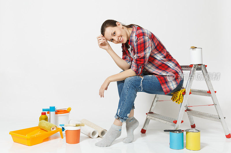 漂亮的女人在休闲衣服坐在梯子与仪器装修公寓房间隔离在白色的背景。墙纸，胶水配件，油漆工具。修理家里的概念。