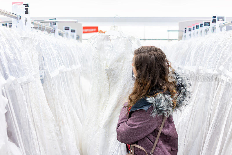 年轻女子拿着蕾丝婚纱在精品店折扣店，许多白色服装挂在衣架排