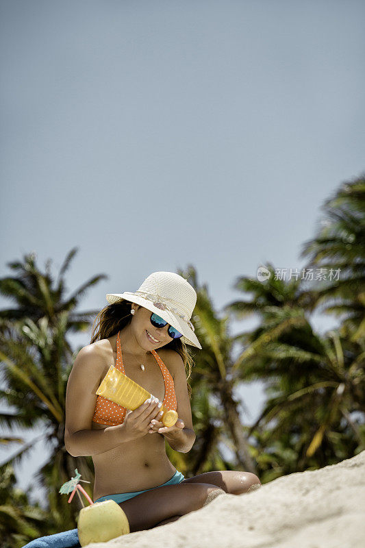 一个西班牙裔年轻女子的特写，在加勒比海的一个热带白沙岛海滩上晒太阳和涂防晒霜