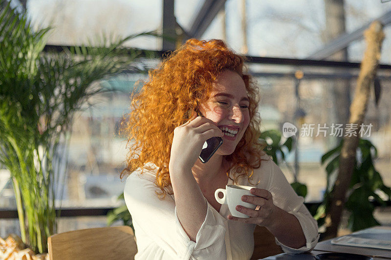 漂亮的红发年轻女子在咖啡馆里讲智能手机