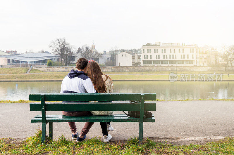 一对年轻夫妇坐在河边的木凳上。美丽的阳光灿烂的日子。背影深情的家庭。快乐的年轻人在公园户外放松