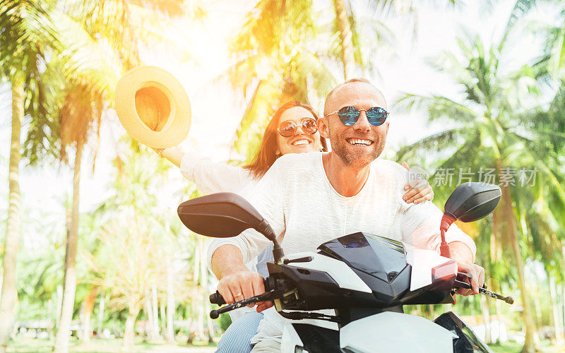一对快乐的旅行者在泰国的热带度假棕榈树下骑摩托车。妇女举着帽子。